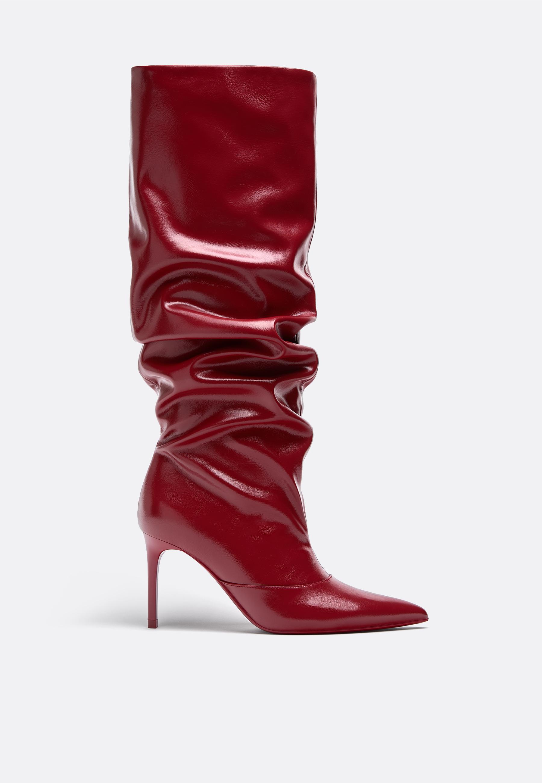 Side view of a red high-heeled boot with a stiletto heel and pointed toe, with a ruched design at the top.