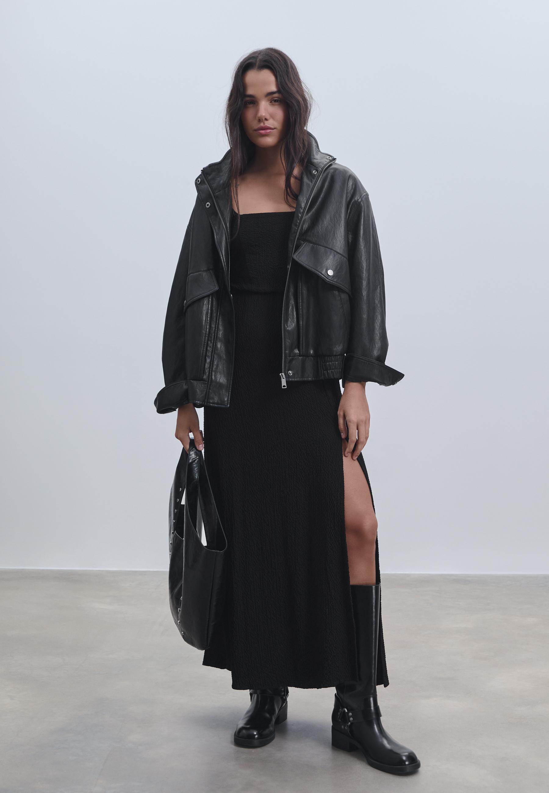 Long black dress set with crinkled texture, square neckline, side slit, black jacket, and tall boots.